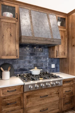 Custom RTM home kitchen with gas cooktop, rustic wood cabinetry, blue tile backsplash, and a farmhouse-style vent hood.