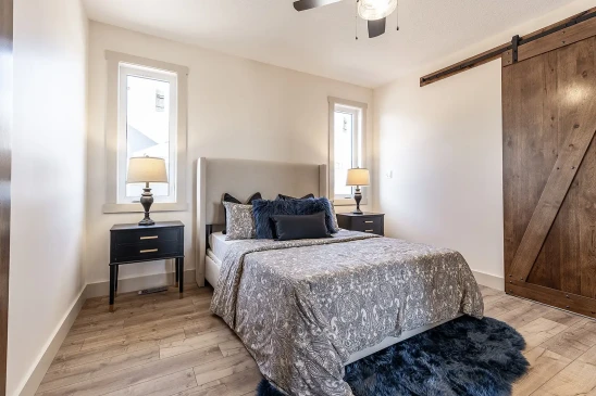Cozy bedroom in a custom RTM home with wood barn door, modern ceiling fan, grey patterned bedding, and natural light from tall windows.