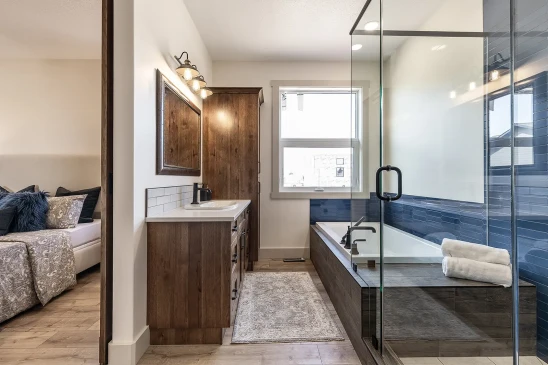 Modern ensuite bathroom with glass shower, deep soaking tub with blue tile accent, wood vanity, and view into adjoining bedroom.