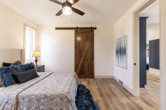 Bright bedroom with modern ceiling fan, wood barn door, and grey patterned bedding accented with blue throw pillows and rug.