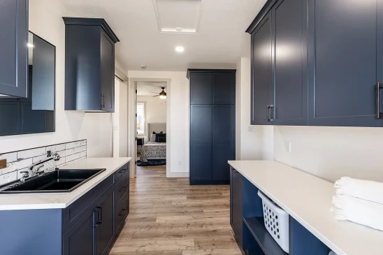 Spacious laundry room with navy blue cabinets, black sink, white countertops, and wood-look flooring leading to a bedroom.