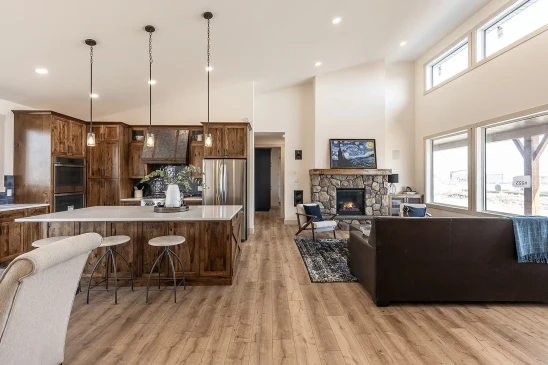 Open-concept RTM home interior featuring a rustic kitchen with wood cabinetry, large island with pendant lighting, and a cozy living room with stone fireplace and expansive windows.