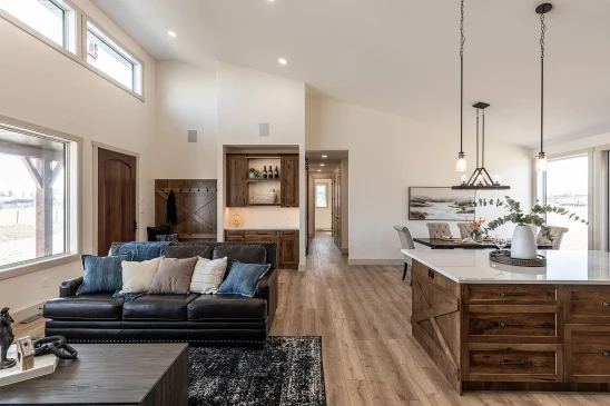 Bright open-concept RTM home interior with wood accents, featuring a modern kitchen island with pendant lighting, cozy living room seating, built-in bar, and dining area with large windows.