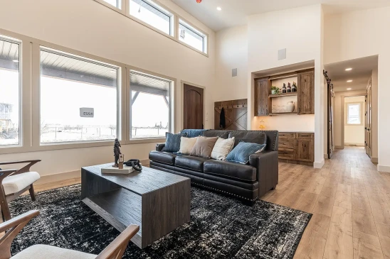 Bright RTM home living room with large energy-efficient windows, black leather sofa, rustic wood cabinetry, and open-concept design with natural light.