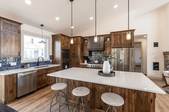 Modern farmhouse kitchen with natural wood cabinetry, quartz island with bar seating, stainless steel appliances, pendant lighting, and tiled backsplash.