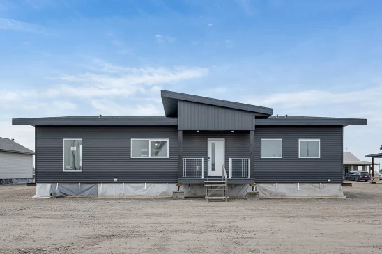 A modern ready-to-move (RTM) home with sleek gray siding and a contemporary flat roof design. The home features a small front porch with white railings and a central front door, accessed by a temporary set of stairs. The exterior includes several large windows that allow natural light to enter the interior. The home is elevated, resting on supports and covered with protective material at the base, indicating it is in the final stages before transport. Surrounding the home are other RTM homes in various stages of construction, and a "Sold" sign is visible next to one of the homes. The sky is partly cloudy, creating a neutral backdrop for the construction site.