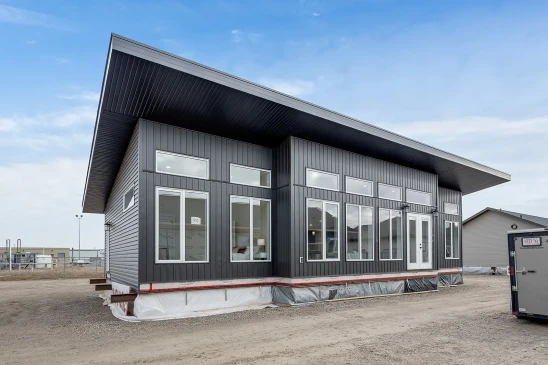 A modern ready-to-move (RTM) home with sleek gray siding and a contemporary flat roof design. The home features a small front porch with white railings and a central front door, accessed by a temporary set of stairs. The exterior includes several large windows that allow natural light to enter the interior. The home is elevated, resting on supports and covered with protective material at the base, indicating it is in the final stages before transport. Surrounding the home are other RTM homes in various stages of construction, and a "Sold" sign is visible next to one of the homes. The sky is partly cloudy, creating a neutral backdrop for the construction site.