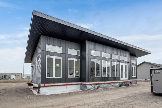 A modern ready-to-move (RTM) home with sleek gray siding and a contemporary flat roof design. The home features a small front porch with white railings and a central front door, accessed by a temporary set of stairs. The exterior includes several large windows that allow natural light to enter the interior. The home is elevated, resting on supports and covered with protective material at the base, indicating it is in the final stages before transport. Surrounding the home are other RTM homes in various stages of construction, and a "Sold" sign is visible next to one of the homes. The sky is partly cloudy, creating a neutral backdrop for the construction site.