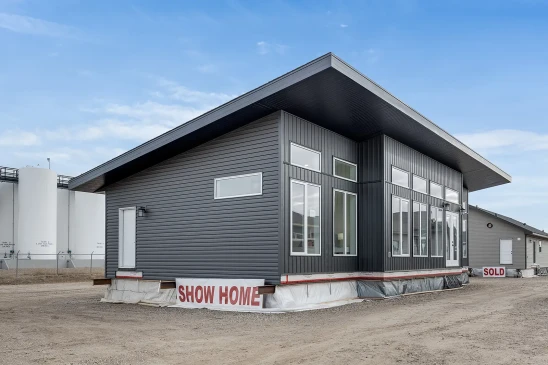 A modern ready-to-move (RTM) home with sleek gray siding and a contemporary flat roof design. The home features a small front porch with white railings and a central front door, accessed by a temporary set of stairs. The exterior includes several large windows that allow natural light to enter the interior. The home is elevated, resting on supports and covered with protective material at the base, indicating it is in the final stages before transport. Surrounding the home are other RTM homes in various stages of construction, and a "Sold" sign is visible next to one of the homes. The sky is partly cloudy, creating a neutral backdrop for the construction site.
