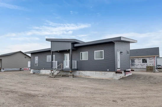 A modern ready-to-move (RTM) home with sleek gray siding and a contemporary flat roof design. The home features a small front porch with white railings and a central front door, accessed by a temporary set of stairs. The exterior includes several large windows that allow natural light to enter the interior. The home is elevated, resting on supports and covered with protective material at the base, indicating it is in the final stages before transport. Surrounding the home are other RTM homes in various stages of construction, and a "Sold" sign is visible next to one of the homes. The sky is partly cloudy, creating a neutral backdrop for the construction site.