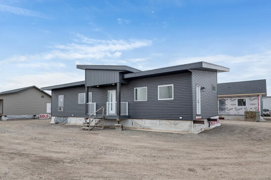 A modern ready-to-move (RTM) home with sleek gray siding and a contemporary flat roof design. The home features a small front porch with white railings and a central front door, accessed by a temporary set of stairs. The exterior includes several large windows that allow natural light to enter the interior. The home is elevated, resting on supports and covered with protective material at the base, indicating it is in the final stages before transport. Surrounding the home are other RTM homes in various stages of construction, and a "Sold" sign is visible next to one of the homes. The sky is partly cloudy, creating a neutral backdrop for the construction site.