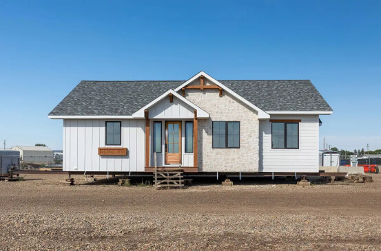 A single-level, ready-to-move (RTM) home with a modern farmhouse exterior. The home features a combination of white siding, stone accents, and wooden trim around the windows and entrance. The front door has a wooden finish with glass panes, and the home sits elevated on a lot, ready for transport to its permanent location. The setting is on a construction lot with clear blue skies overhead.