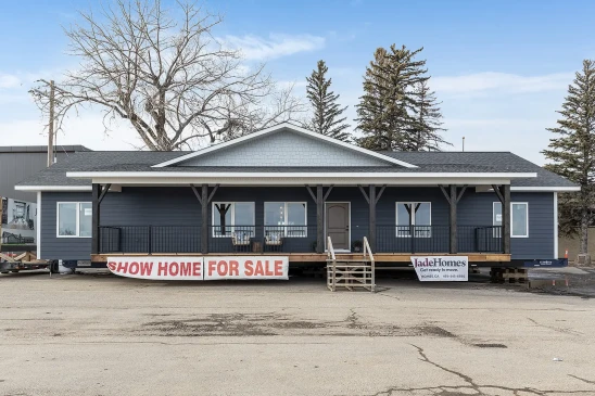 A dark blue ready-to-move (RTM) show home with a large front porch, displayed for sale on a lot. The home features a wide, single-story layout with dark trim, white accents, and a gabled roof. A banner at the front reads "Show Home For Sale," and stairs lead up to the entrance. The setting is an open area with trees and industrial buildings in the background, showing the home in its transportable stage.