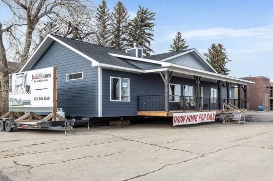 Dark blue ready-to-move (RTM) show home with a large front porch, displayed for sale on a lot. The home features a wide, single-story layout with dark trim, white accents, and a gabled roof. A banner at the front reads "Show Home For Sale," and stairs lead up to the entrance. The setting is an open area with trees and industrial buildings in the background, showing the home in its transportable stage.