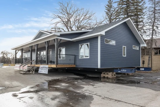 A dark blue ready-to-move (RTM) show home with a large front porch, displayed for sale on a lot. The home features a wide, single-story layout with dark trim, white accents, and a gabled roof. A banner at the front reads "Show Home For Sale," and stairs lead up to the entrance.