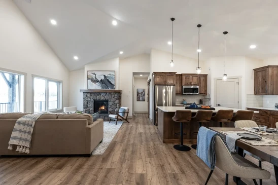 A spacious and modern interior of a ready-to-move (RTM) home featuring an open-concept living room and kitchen. The living area includes a cozy stone fireplace, beige sectional sofa, and large windows that allow natural light to flood the space. The kitchen is equipped with wooden cabinets, stainless steel appliances, an island with bar seating, and pendant lighting. A dining table is set nearby, completing the warm and inviting atmosphere of this stylish home. The hardwood flooring and vaulted ceiling add to the home's elegant yet comfortable design.