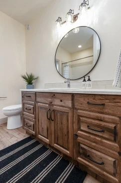 An inviting bathroom showcasing a rustic wooden vanity with ample storage, paired with a round mirror framed in black for a modern touch. The vanity is topped with a sleek white countertop and complemented by elegant lighting fixtures above. A nearby potted plant adds a fresh, natural element to the design, creating a warm and functional space.