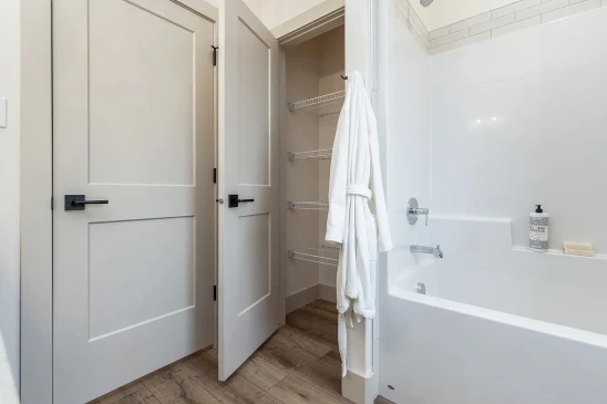 A modern bathroom featuring a white bathtub with a sleek chrome faucet and showerhead, accompanied by a linen closet with wire shelving for organized storage. The space includes minimalistic paneled doors with black hardware and a cozy white robe hanging nearby, creating a clean and inviting atmosphere.