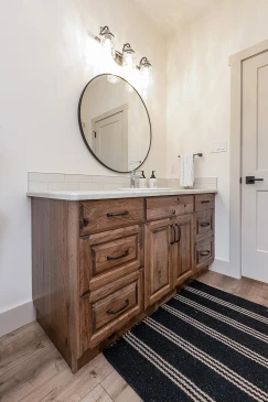 A stylish bathroom vanity featuring a rich wood cabinet with black hardware, topped with a quartz countertop and a round mirror. The space is illuminated by elegant glass light fixtures, while a striped area rug adds a touch of texture to the wooden flooring.