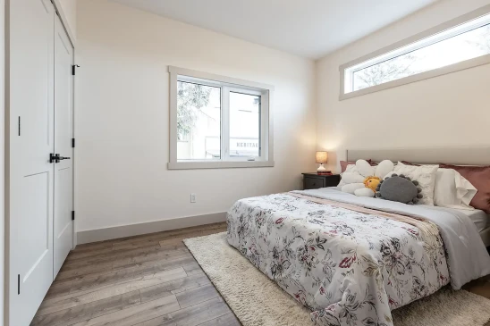 A serene bedroom featuring a plush bed with floral bedding, accented by decorative pillows, including a flower-shaped cushion. The room is illuminated by natural light from a horizontal window and two bedside lamps on matching dark nightstands, with warm wooden flooring and a cozy area rug completing the space.