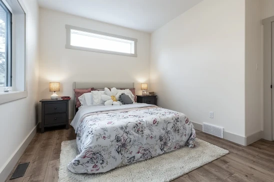 A serene bedroom featuring a plush bed with floral bedding, accented by decorative pillows, including a flower-shaped cushion. The room is illuminated by natural light from a horizontal window and two bedside lamps on matching dark nightstands, with warm wooden flooring and a cozy area rug completing the space.