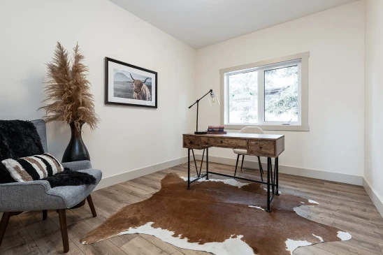 A cozy home office with a rustic wooden desk, a comfortable chair, and a cowhide rug on wood-style flooring. The room is decorated with a framed photograph of a Highland cow, a vase with dried pampas grass, and a plush armchair featuring decorative pillows for added warmth and style.