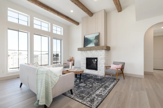 Open and airy living room with large floor-to-ceiling windows, a stone fireplace with a wood mantel, exposed wooden ceiling beams, and cozy seating featuring a soft sofa and modern armchairs, creating a blend of rustic and contemporary design.