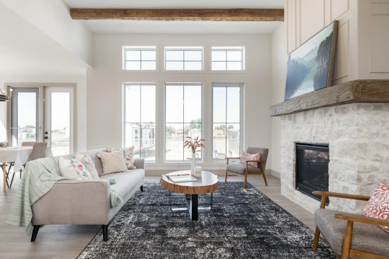 A bright and modern living room featuring a large stone fireplace with a wooden mantel, topped by a flat-screen TV. The space is decorated with neutral tones and floral accent pillows on a light gray sofa. The seating area also includes a pair of mid-century modern armchairs and a wooden coffee table set on a textured black and white area rug. The room is filled with natural light from large, grid-patterned windows, and an exposed wooden beam adds a rustic touch to the otherwise contemporary design. The open-concept layout extends to a dining area in the background, creating a welcoming and airy atmosphere.