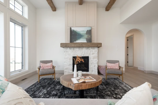 Cozy living room with a rustic stone fireplace topped by a natural wood mantel, framed by exposed wood beams. The space features modern armchairs with pink accent pillows, a live-edge coffee table, and large windows allowing abundant natural light.