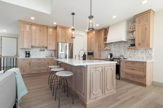 Spacious and inviting farmhouse kitchen featuring natural wood cabinetry, a large island with intricate paneling, white countertops, brass fixtures, pendant lighting, stainless steel appliances, and a stone backsplash complemented by open shelving.