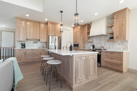 Spacious and inviting farmhouse kitchen featuring natural wood cabinetry, a large island with intricate paneling, white countertops, brass fixtures, pendant lighting, stainless steel appliances, and a stone backsplash complemented by open shelving.