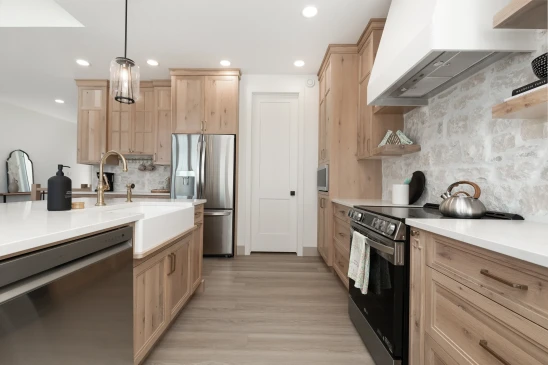 Modern farmhouse kitchen in a RTM Home featuring light wood cabinets, a white farmhouse sink with a brass faucet, stainless steel appliances, a stone backsplash, open shelving, and a spacious island with pendant lighting.