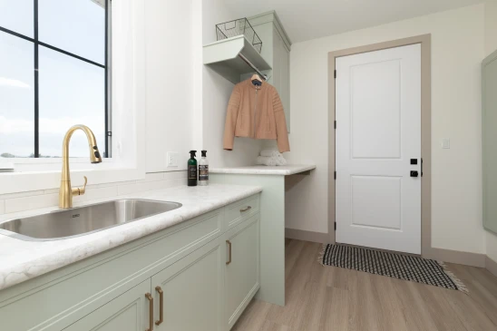 Bright and functional laundry room featuring mint green cabinets with brass hardware, a stainless steel sink with a gold faucet, a folding station with hanging space, and a modern design accented by natural light from a large window.