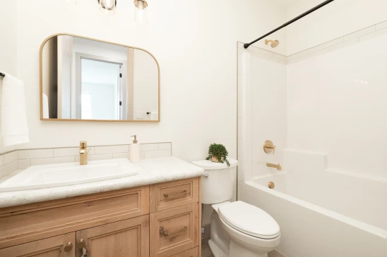 Modern bathroom with a light wood vanity, white marble countertop, gold fixtures, a sleek arched mirror, and a clean white bathtub with a black shower curtain rod, creating a bright and elegant design.