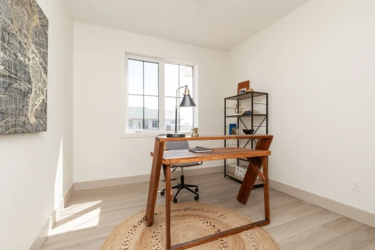 A bright, minimalist home office featuring a wooden desk with a modern, angular design. The room is illuminated by natural light from a large window with white trim. A black industrial-style desk lamp adds a functional yet stylish touch to the space. Behind the desk is a black metal shelving unit with open shelves, decorated with books, ornaments, and a small photo frame. A round woven rug adds warmth and texture to the light wood flooring. A piece of abstract art is mounted on the wall, giving the room a subtle artistic flair.