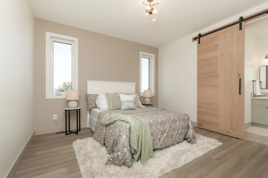 A cozy bedroom with a neutral color palette featuring a queen-sized bed with a white headboard and gray paisley-patterned bedding. A light green throw blanket adds a soft accent to the bed. The room has two tall, narrow windows on either side of the bed, allowing natural light to brighten the space. On each side of the bed are round bedside tables with matching lamps. A sliding wooden barn door, leading to an en suite bathroom, adds a rustic touch to the modern design. The room is finished with light wood flooring and a plush white area rug under the bed.