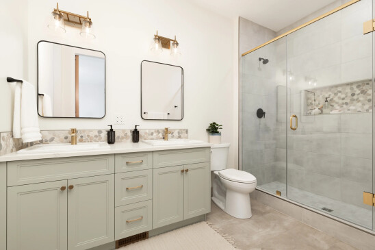 A modern and elegant bathroom featuring a double vanity with light gray cabinetry, gold hardware, and two square mirrors with black frames. Above each mirror is a brass light fixture with glass shades, adding a touch of sophistication. The countertop is white with a pebble-patterned backsplash. To the right is a large glass-enclosed shower with a gold handle and black fixtures, complemented by gray tile walls and a built-in shelf for toiletries. The bathroom is bright, with neutral tones, creating a clean and inviting space.