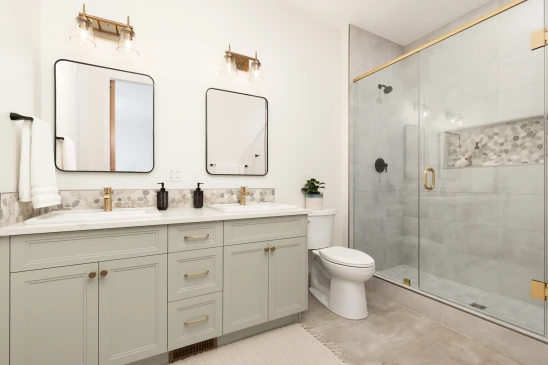 A modern and elegant bathroom featuring a double vanity with light gray cabinetry, gold hardware, and two square mirrors with black frames. Above each mirror is a brass light fixture with glass shades, adding a touch of sophistication. The countertop is white with a pebble-patterned backsplash. To the right is a large glass-enclosed shower with a gold handle and black fixtures, complemented by gray tile walls and a built-in shelf for toiletries. The bathroom is bright, with neutral tones, creating a clean and inviting space.